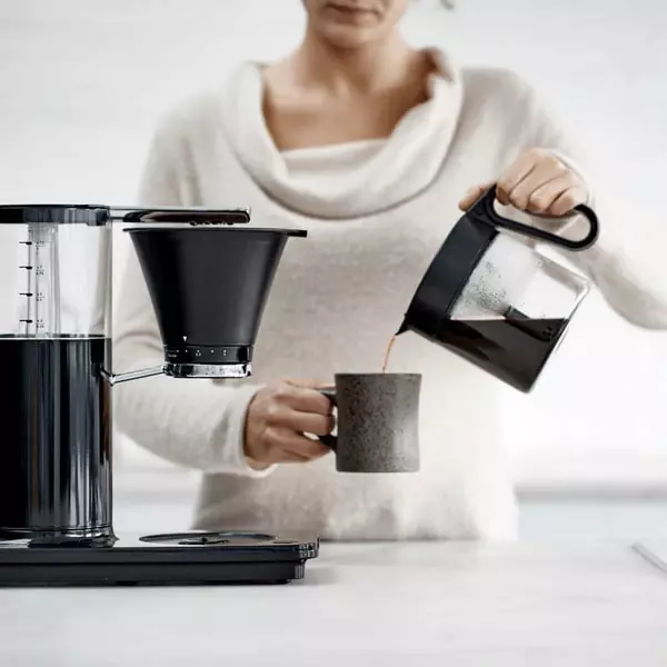Dụng cụ nhỏ giọt Wilfa Classic+ và một người phụ nữ đang rót cà phê từ ấm đun nước vào cốc.